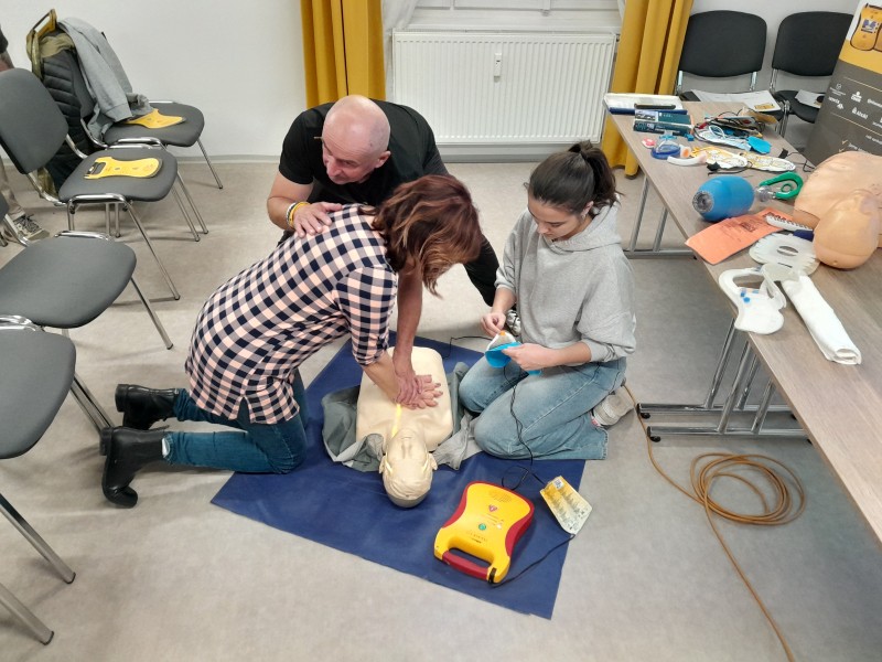 Na obrázku dva občané provádějí nácvik masáže srdce a používání externího defibrilátoru pod dohledem lektora.