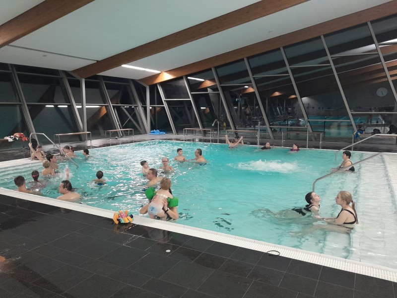 Na fotografii jsou děti i dospělí v bazénu, plavou a baví se.